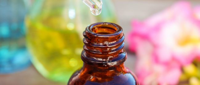 A clear glass eyedropper adds some drops of liquid to an open brown glass bottle. Flowers and glass perfume bottles are visible in the background.