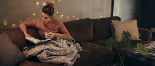 A woman reads a book on her couch under a blanket. She is wearing a sweater and drinking a warm drink. The room is dimly lit with a string of lights.