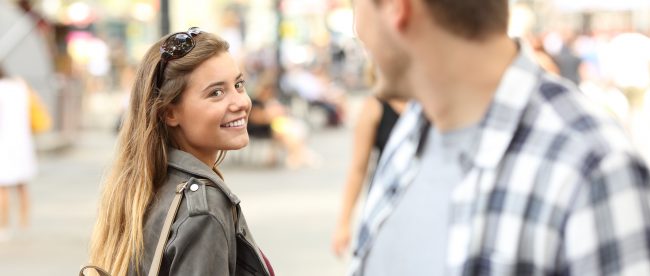 A happy woman looks over her shoulder and smiles at a man she is passing on the street.