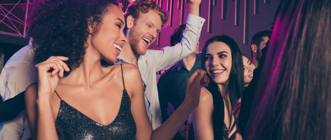 Friends dancing and having fun on the dance floor of a club. Two women are smiling and laughing at each other while the man behind them raises one hand in the air in excitement.
