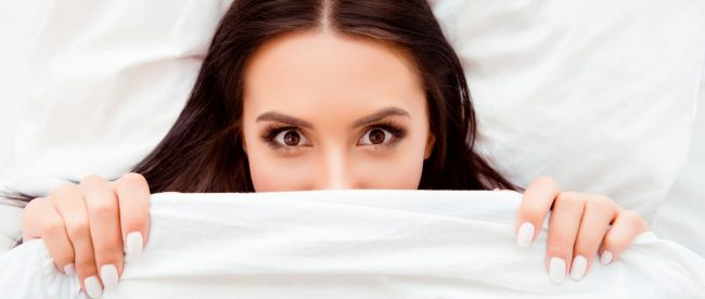 Smiling woman lying on her back in bed with the covers pulled up to cover half her face.