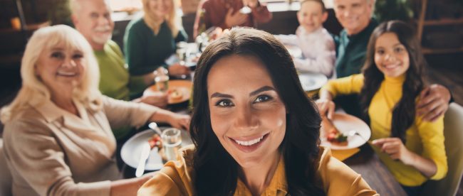 A woman takes a selfie with her family in the background. They are sitting down to a big family holiday dinner.