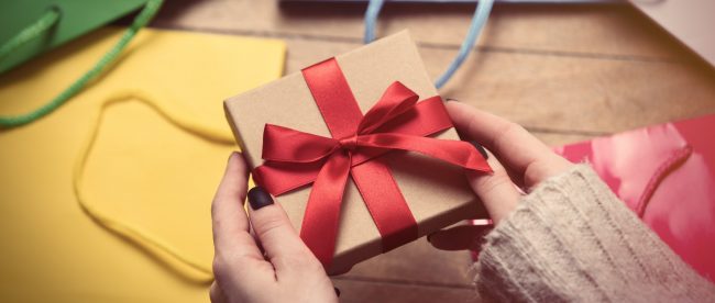 A close-up view of a woman's hands holding a wrapped gift box. Several gift bags are spread out on the floor in front of her.