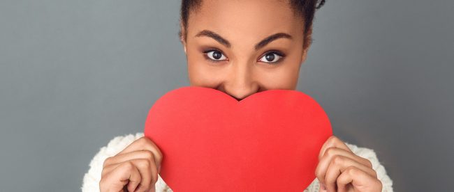 A smiling woman holds a paper heart up to cover the lower half of her face.