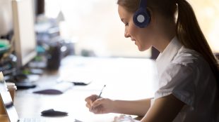 A woman works at a desk in an office. She is wearing headphones and writing on a notebook in front of her tablet.