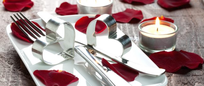 A romantic place setting on a wooden table: tea candles, and rose petals surround a small square plate that holds silverware and heart-shaped napkin rings.