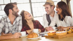 Four friends, two men and two women, sit in a coffee shop and laugh together. They have drinks and pastries in front of them.