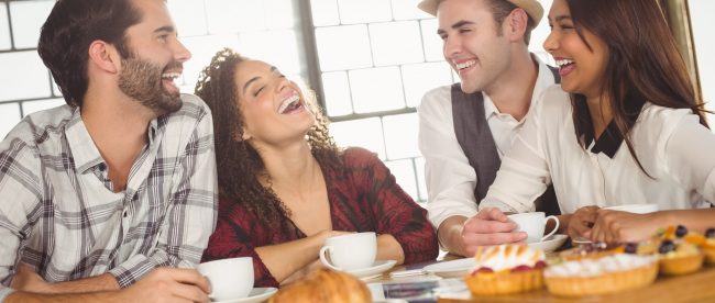 Four friends, two men and two women, sit in a coffee shop and laugh together. They have drinks and pastries in front of them.