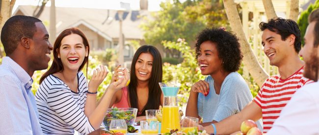 A group of smiling friends sit at a table outdoors eating lunch.