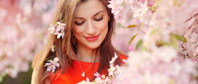 A smiling woman in a red dress is standing very close to a flowering tree and is surrounded by pink blossoms.