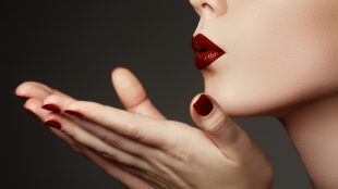 A close up view of a woman wearing red lipstick and red nail polish blowing a kiss