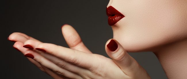 A close up view of a woman wearing red lipstick and red nail polish blowing a kiss