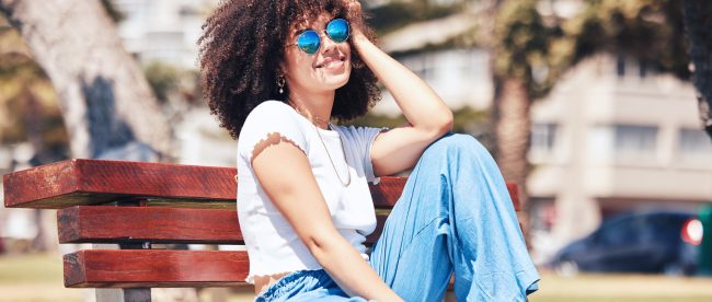 A woman with curly hair, sunglasses, a plain white tee shirt and blue jeans smiles in the sunshine on a city park bench.