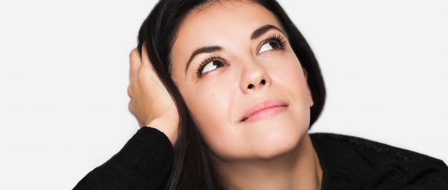 A brown-haired woman is thinking about something with her head rested in her hand and her eyes pointed upwards. She is smiling.