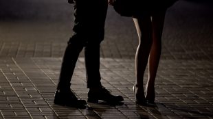 A man and woman are standing on a paved brick street at nighttime. Only their legs are visible. The man is wearing a suit and lace-up shoes and the woman is wearing a short tress with high-heeled shoes.