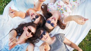 Four women in summer clothes and suglasses laying on their back on a white sheet in a grassy field. They are all happy and smiling and pointing up at the camera.