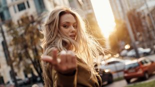 A woman with messy blonde hair beckons toward the camera as she walks down a busy city avenue at sunset.
