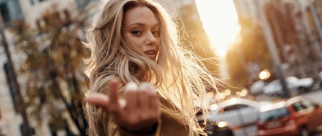 A woman with messy blonde hair beckons toward the camera as she walks down a busy city avenue at sunset.