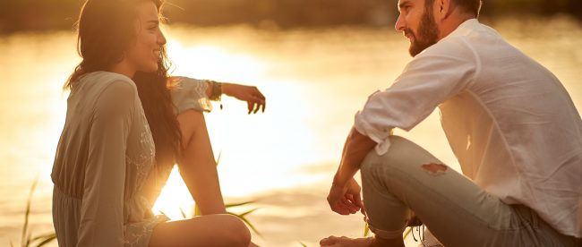 A young couple sits on a dock at sunset, facing each other and smiling, having a pleasant conversation