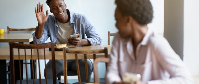 A woman sitting in a cafe looks over her shoulder to see a happy man smiling and waving back at her.