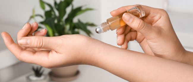 A photo of a woman's hands applying a roll-on bottle of oil to her wrist over a bathroom counter. A potted plant and bottle of perfume sit in the background.