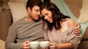 A smiling man and woman in long sleeved shirts sit on the floor with their backs against their couch. They are drinking hot cocoa and leaning into one another with their heads pressed together.