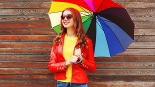 A smiling woman in a red jacket and red heart-shaped sunglasses holds a rainbow-colored umbrella in front of a wood-paneled wall.