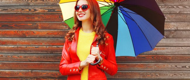 A smiling woman in a red jacket and red heart-shaped sunglasses holds a rainbow-colored umbrella in front of a wood-paneled wall.