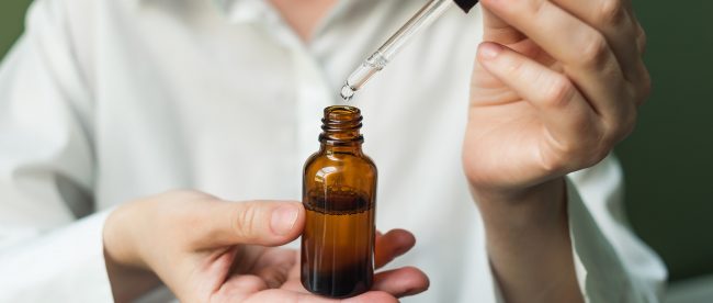 A woman in a white shirt uses an eyedropper to add drops of liquids to a brown glass bottle that she is holding in her hands.