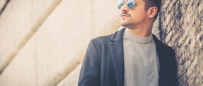 A masculine man with stubble and sunglasses leans against a brick wall.