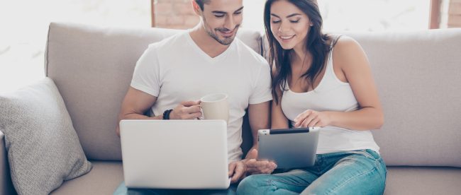 A man and woman sit on a couch close together. He has a laptop in his lap, and she has a tablet in her hands. They both look at her tablet with smiles on their faces.