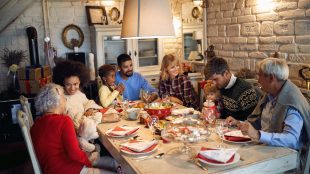 A multiracial family of six adults and two children sit around a holiday table. The table is covered in cookies and place settings with cloth napkins. Everyone is talking together and enjoying each other's company.