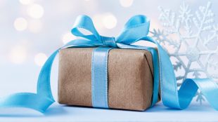A gift wrapped in brown paper wth a light blue ribbon and bow sits on a table wnext to snowflakes. Winter lights are in the background.