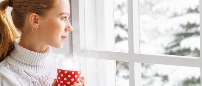 A young woman wearing a white turtleneck and holding a red and white polka dot mug looks out of her window onto a snowy winter landscape