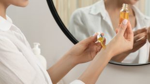 A woman applies golden oil to her wrists in front of a mirror.