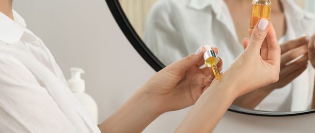 A woman applies golden oil to her wrists in front of a mirror.