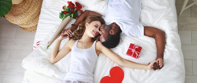 OVerhead view of a man and woman snuggling in bed. They are lying face-to-face, holding hands, and a bouquet of roses and wrapped gift package are on the bed with them.
