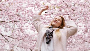 A woman in a sweater and scarf stretches her arms up and smiles underneath a canopy of blooming trees in springtime.