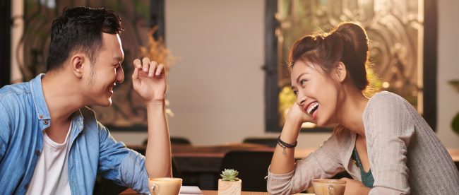 A man and woman sit at a table in a coffee shop, leaning towards each other and laughing happily.