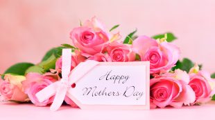 A bouquet of pink roses lays sideways on a table in front of a soft pink background. In front of them, a gift tag reads "Happy Mother's Day."