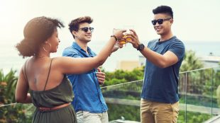 Two young men and a young woman stand on an outdoor balcony and clink their glasses together in a toast.