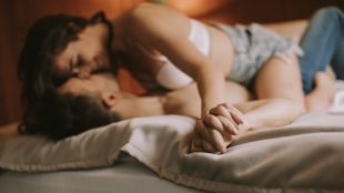 A woman straddles a man in their bed. She is in a bra and shorts and he is in jeans. Neither are wearing a shirt. Their fingers are intertwined and they are both smiling.