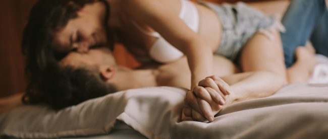 A woman straddles a man in their bed. She is in a bra and shorts and he is in jeans. Neither are wearing a shirt. Their fingers are intertwined and they are both smiling.
