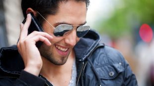 A man in sunglasses and black jacket smiles while talking on his cell phone in the street