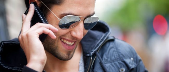 A man in sunglasses and black jacket smiles while talking on his cell phone in the street