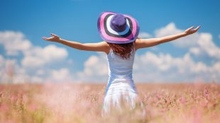 A woman stands in a field of flowers in summertime. Her back is to the camera and her arms are spread wide. She is in a sundress and sun hat. The sky is blue with white clouds on the horizon.