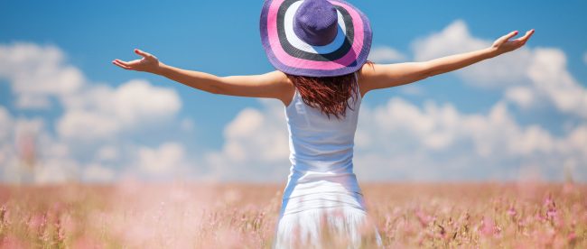 A woman stands in a field of flowers in summertime. Her back is to the camera and her arms are spread wide. She is in a sundress and sun hat. The sky is blue with white clouds on the horizon.